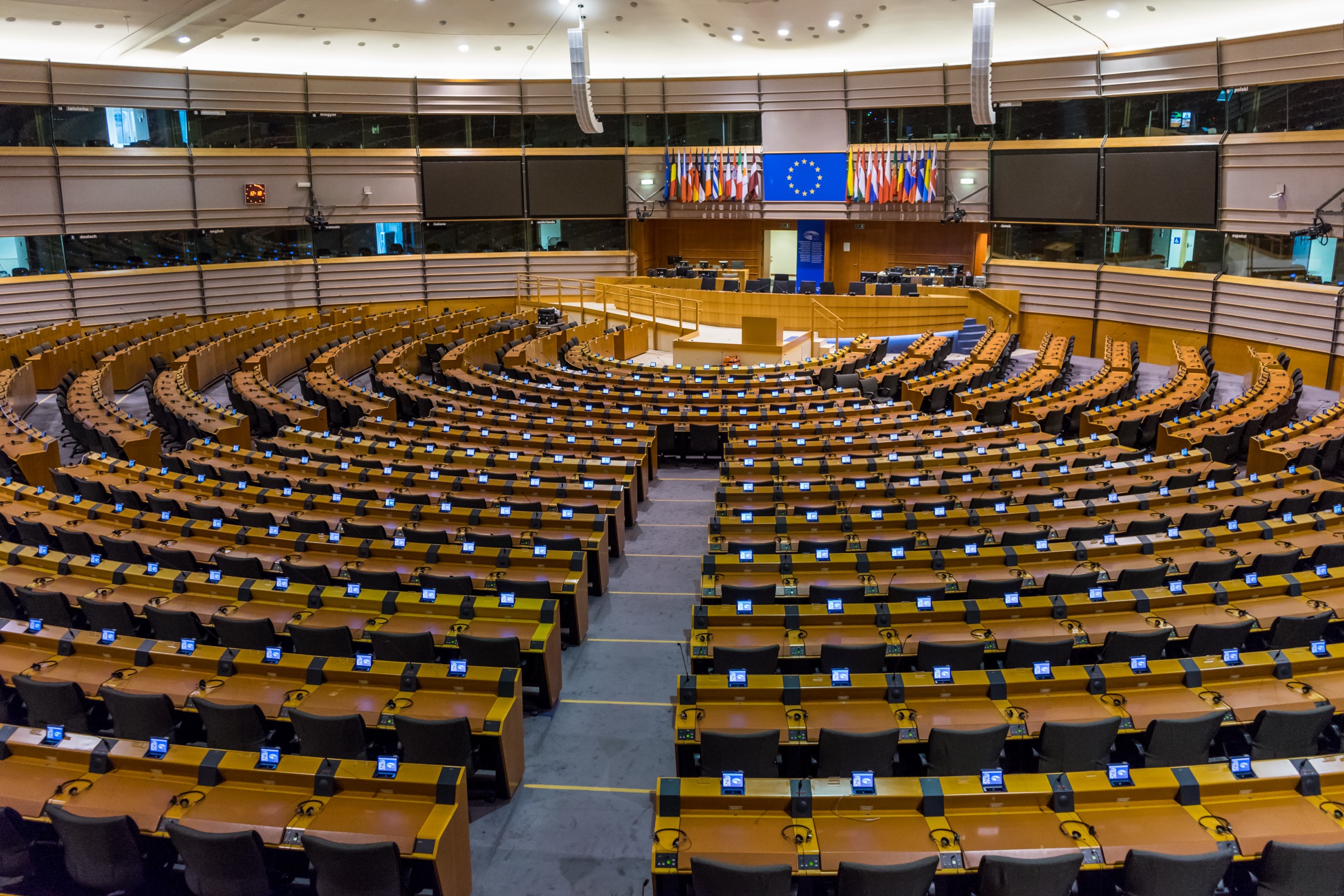European Parliament empty plenary chamber, Brussels — curved tiers of member seats, the central speaker tribune, and the EU flag rotunda above